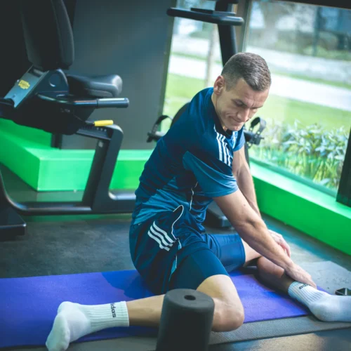 Mężczyzna w niebieskim stroju sportowym rozciąga nogi na macie w siłowni, obok sprzętu do ćwiczeń.