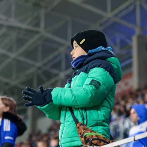 Na zdjęciu widać chłopca w zielonej kurtce, który w fanatyczny sposób kibicuje z trybun na stadionie.