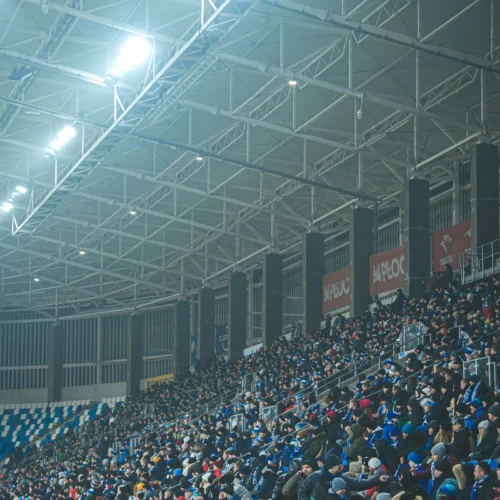 Na zdjęciu widać zatłoczone trybuny stadionu, pełne kibiców ubranych w różne odcienie niebieskiego i czarnego, pod jasnym oświetleniem.