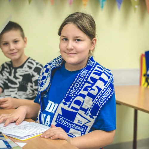 Na zdjęciu widać dwoje dzieci siedzących przy biurku w jasnej sali, z uśmiechem i przyborami szkolnymi, gdzie jedno z nich trzyma kartkę, a drugie ma na sobie niebieską koszulkę oraz szalik.