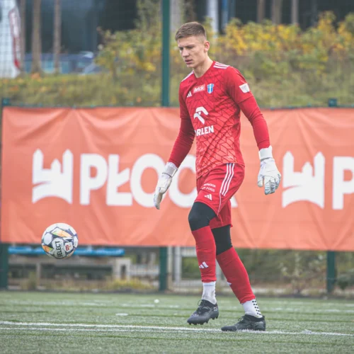 Na zdjęciu widać bramkarza w czerwonym stroju, który stoi na boisku piłkarskim, gotowy do interwencji, z piłką uniesioną w powietrzu.