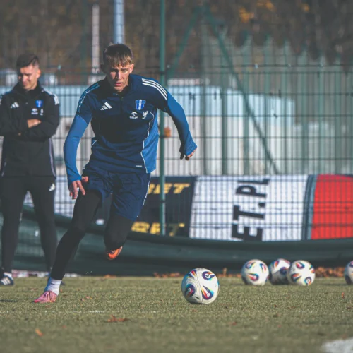 Na zdjęciu widać młodego piłkarza w niebieskim stroju, który biegnie w kierunku piłek na boisku, podczas gdy drugi zawodnik obserwuje jego trening.