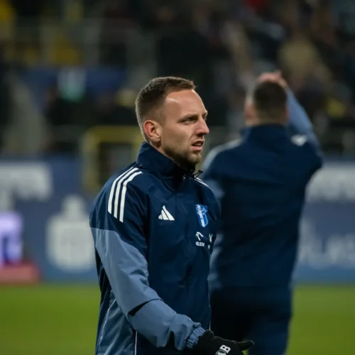 Zdjęcie przedstawia mężczyznę w granatowej kurtce sportowej, który obserwuje boisko piłkarskie, podczas gdy w tle widać innych ludzi na stadionie.