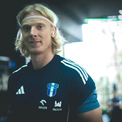 Na zdjęciu widać młodego mężczyznę z długimi włosami, w sportowym stroju z logo klubu, uśmiechającego się w jasnym otoczeniu.