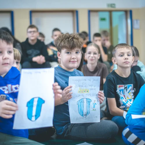 Grupa dzieci siedzi na podłodze, trzymając w rękach swoje rysunki i uśmiechając się w trakcie zajęć.