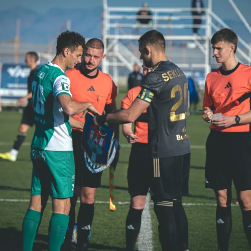 Na zdjęciu widać zawodników drużyn piłkarskich wymieniających się klubowymi szalami przed rozpoczęciem meczu, a także sędziów przygotowujących się do spotkania.