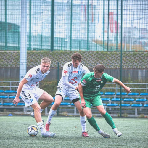 Na zdjęciu widoczni są trzej młodzi piłkarze walczący o piłkę na boisku, przy ogrodzeniu i trybunach w tle.