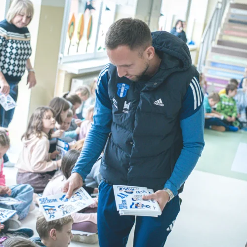 Na zdjęciu widoczny jest mężczyzna rozdający dzieciom materiały, podczas gdy uczniowie siedzą na podłodze w sali lekcyjnej.