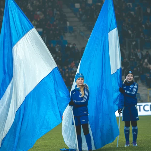 Na zdjęciu widać dwie dziewczynki w niebieskich strojach sportowych, które trzymają duże, niebiesko-białe flagi na boisku podczas wydarzenia sportowego.