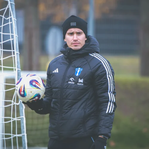 Mężczyzna w czarnej kurtce i czapce trzyma piłkę nożną obok bramki na boisku treningowym.