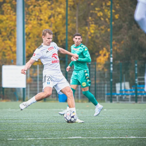 Na zdjęciu widoczny jest piłkarz w białym stroju, który wykonuje kopnięcie piłki na boisku, z bramkarzem w zielonym stroju w tle.