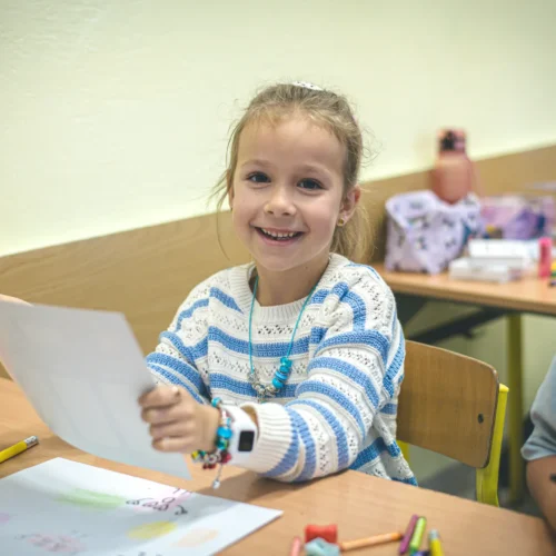 Uśmiechnięta dziewczynka trzyma kartkę w rękach, z kolorowymi przyborami plastycznymi rozłożonymi na stole w tle.