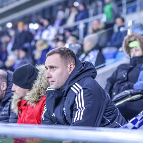 Na zdjęciu widoczna jest grupa kibiców siedzących na trybunach podczas meczu, skoncentrowanych na wydarzeniach na boisku.