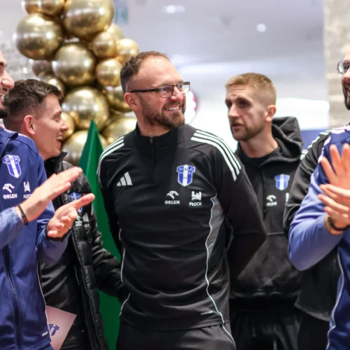 Grupa ludzi w sportowych strojach, uśmiechających się i rozmawiających w towarzystwie złotych dekoracji.