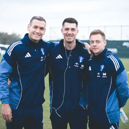 Trzej mężczyźni w niebieskich strojach sportowych uśmiechają się na tle boiska piłkarskiego.