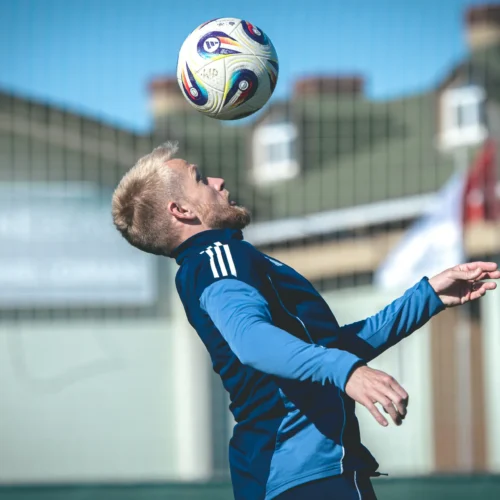 Młody mężczyzna w niebieskim stroju piłkarskim wykonuje ćwiczenie, skacząc, aby zgrać piłkę nad głową na boisku.