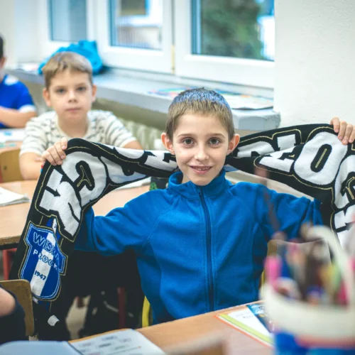 Na zdjęciu widoczny jest uśmiechnięty chłopiec w niebieskiej bluzie, który trzyma w rękach szalik, a w tle siedzi kilku jego kolegów w klasie.