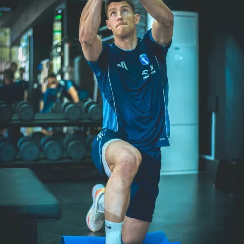 Mężczyzna ćwiczy w siłowni, trzymając ciężar w górze na kolanie, otoczony sprzętem do treningu.