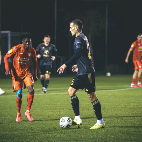 Na zdjęciu widać dwóch piłkarzy podczas meczu, jeden z nich jest w pomarańczowej, a drugi w ciemnej, sportowej koszulce, na tle boiska oświetlonego w nocy.