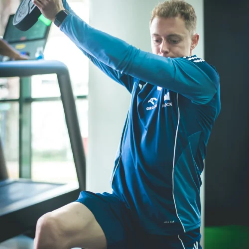 Mężczyzna w niebieskim stroju sportowym wykonuje ćwiczenie siłowe na siłowni, trzymając talerz obciążeniowy nad głową.