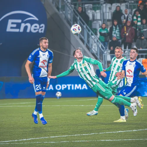 Na zdjęciu widoczni są piłkarze podczas dynamicznej akcji na boisku, z piłką unoszącą się w powietrzu.