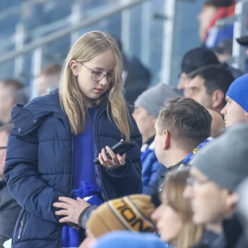 Na zdjęciu widać młodą osobę w niebieskiej kurtce, trzymającą telefon, siedzącą na trybunach podczas meczu, otoczoną innymi kibicami.