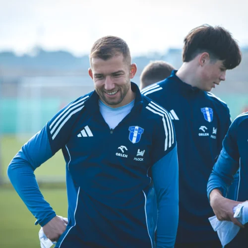 Na zdjęciu trzech zawodników piłkarskich w niebieskich strojach sportowych uśmiecha się podczas treningu na boisku.