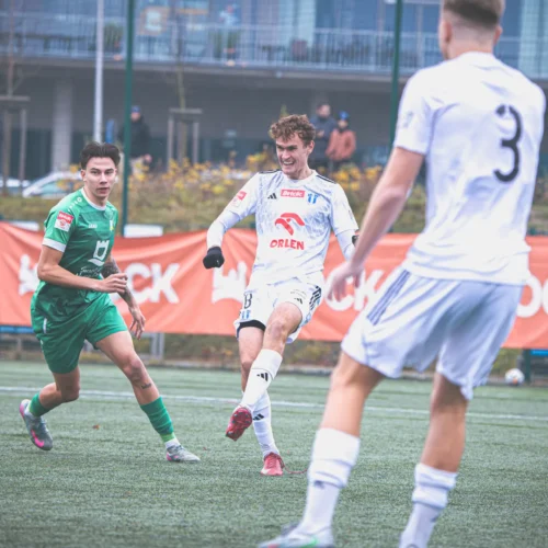 Na zdjęciu zawodnicy podczas meczu piłki nożnej, z jednym z graczy w białym stroju wykonującym kopnięcie piłki, a inni zawodnicy w zielonych strojach obserwują akcję.