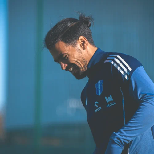 Na zdjęciu znajduje się uśmiechający się mężczyzna w niebieskim stroju sportowym, spacerujący po boisku piłkarskim.