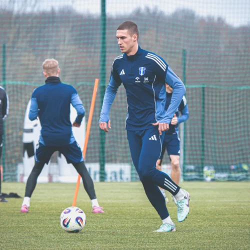 Na zdjęciu widać grupę piłkarzy trenujących na boisku, z jednym zawodnikiem w niebieskim stroju, który biegnie obok piłki.