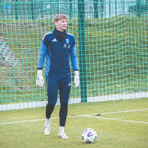 Na zdjęciu widać bramkarza w strój sportowym stojącego na boisku piłkarskim obok piłki.