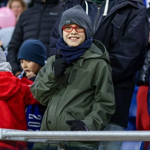 Na zdjęciu widoczny jest uśmiechnięty chłopiec w szarej czapce i okularach, stojący na tle innych kibiców na trybunach stadionu.