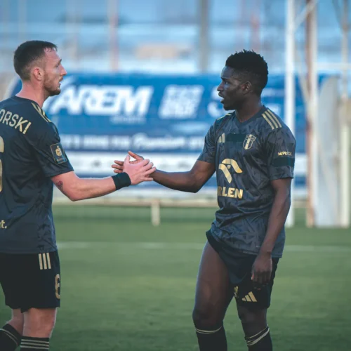 Na zdjęciu dwóch piłkarzy w czarnych strojach z goldowymi detalami uścisnęło sobie dłonie na boisku.