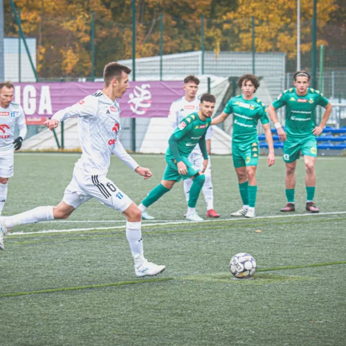 Na boisku piłkarskim zawodnik wykonuje rzut karny, a w tle widoczni są pozostali gracze drużyny przeciwnika.