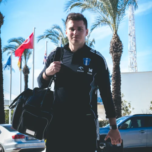 Mężczyzna w sportowym ubraniu niesie torbę oraz walizkę, stojąc na tle palm i flag w słoneczny dzień.