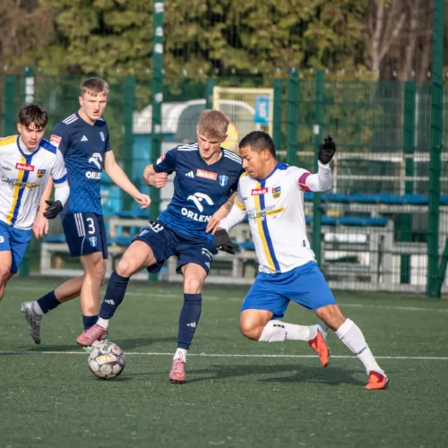 Na zdjęciu widać czterech zawodników footballowych rywalizujących o piłkę na sztucznej trawie.