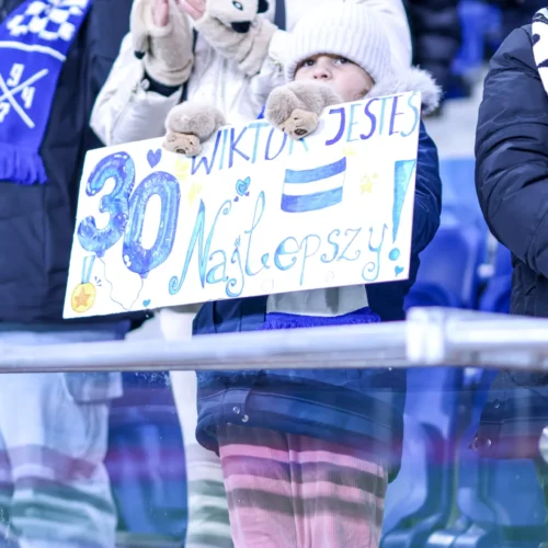 Na zdjęciu widać dziecko w zimowym ubraniu trzymające kolorowy baner z napisem "WIKTOR JESTEŚ NAJLEPSZY!" podczas meczu lub imprezy sportowej.
