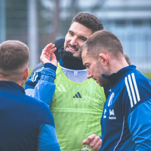 Na zdjęciu trzej mężczyźni w strojnym treningowym rozmawiają ze sobą, z uśmiechami na twarzach, w trakcie treningu piłkarskiego.