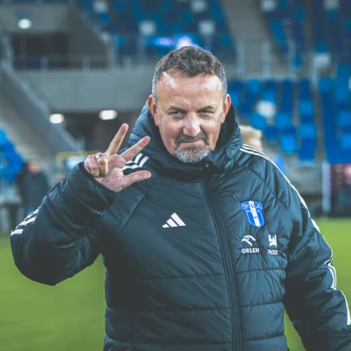 Mężczyzna w czarnej kurtce sportowej stoi na boisku i pokazuje gest „victory” w kierunku kamery.