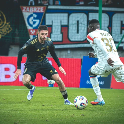 Na zdjęciu dwóch piłkarzy rywalizuje na boisku podczas meczu, z jednym z nich w czarnym stroju i drugim w białym.