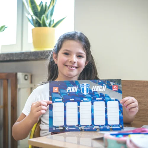 Dziewczynka siedzi przy biurku w świetle dziennym, uśmiechając się i trzymając w rękach planer lekcji z logo Wisły Kraków.