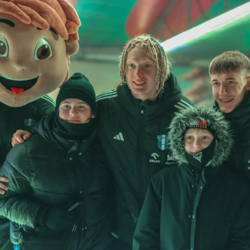 Na zdjęciu widać grupę młodych ludzi w zimowych kurtkach, pozujących z osobą w kostiumie maskotki o uśmiechniętej twarzy.