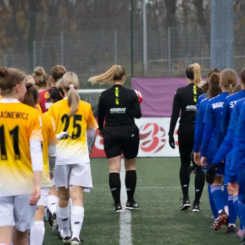 Na zdjęciu widać zawodniczki piłki nożnej w dwóch różnych strojach, które idą na boisku w kierunku sędziów przed rozpoczęciem meczu.
