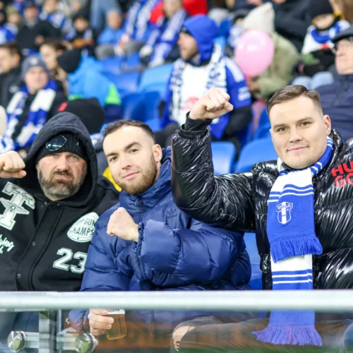 Na zdjęciu widać trzech kibiców, którzy entuzjastycznie pokazują pięści na trybunach stadionu, ubrani w ciepłe kurtki i szaliki w kolorach ich drużyny.