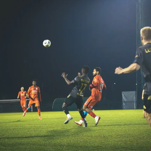 Na zdjęciu widoczna jest akcja w meczu piłkarskim, w której zawodnicy w ciemnych strojach walczą o piłkę z rywalami w pomarańczowych strojach na oświetlonym boisku.