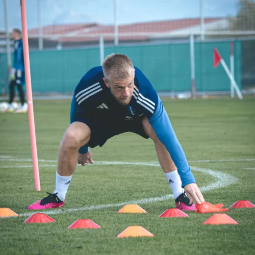 Na zdjęciu widać sportowca wykonującego ćwiczenia na boisku, sprzedającego kolorowe stożki w otoczeniu sprzętu treningowego.