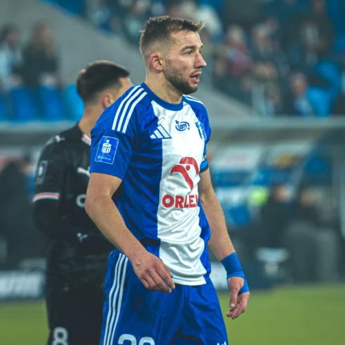 Na zdjęciu widać piłkarza w niebiesko-białym stroju drużyny, stojącego na boisku podczas meczu.