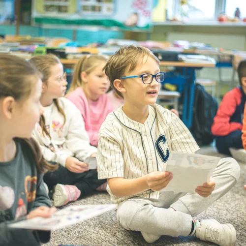 Grupa dzieci siedzi na podłodze w klasie, koncentrując się na zajęciach i trzymając w rękach kartki papieru.