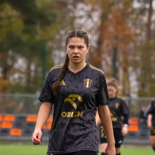 Młoda zawodniczka w czarnej koszulce z logo Orlen idzie w stronę kamery na boisku piłkarskim.