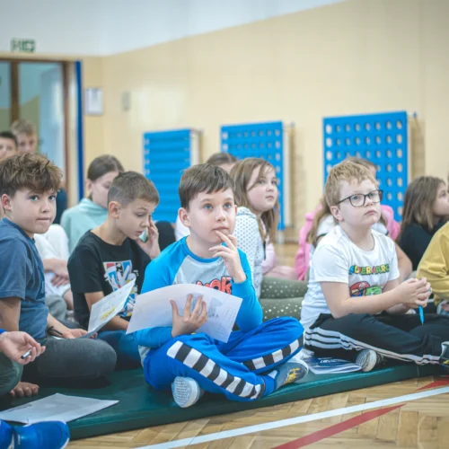 Grupa dzieci siedzi na podłodze, uważnie słuchając, co dzieje się na prowadzonej aktywności.